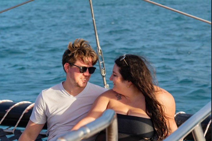 Two people sitting on a sailboat net, smiling and talking over the ocean.