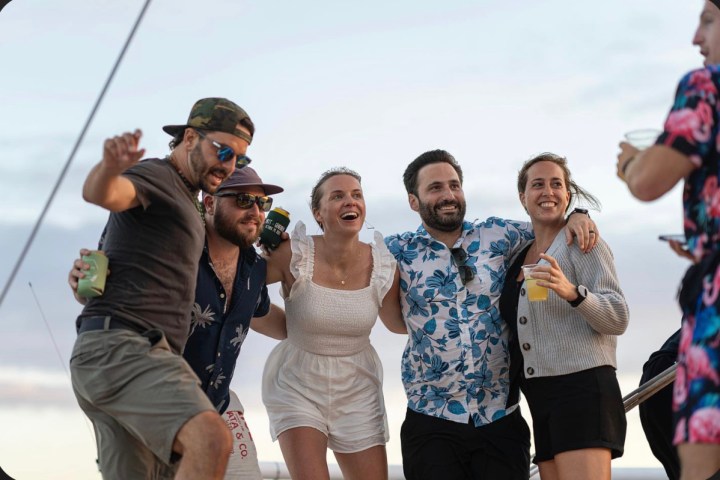 Group of five people smiling on a boat, holding drinks, with one person wearing a floral shirt.